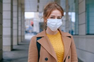 A young red-headed woman with a surgical face mask and a winter coat looks into the camera.