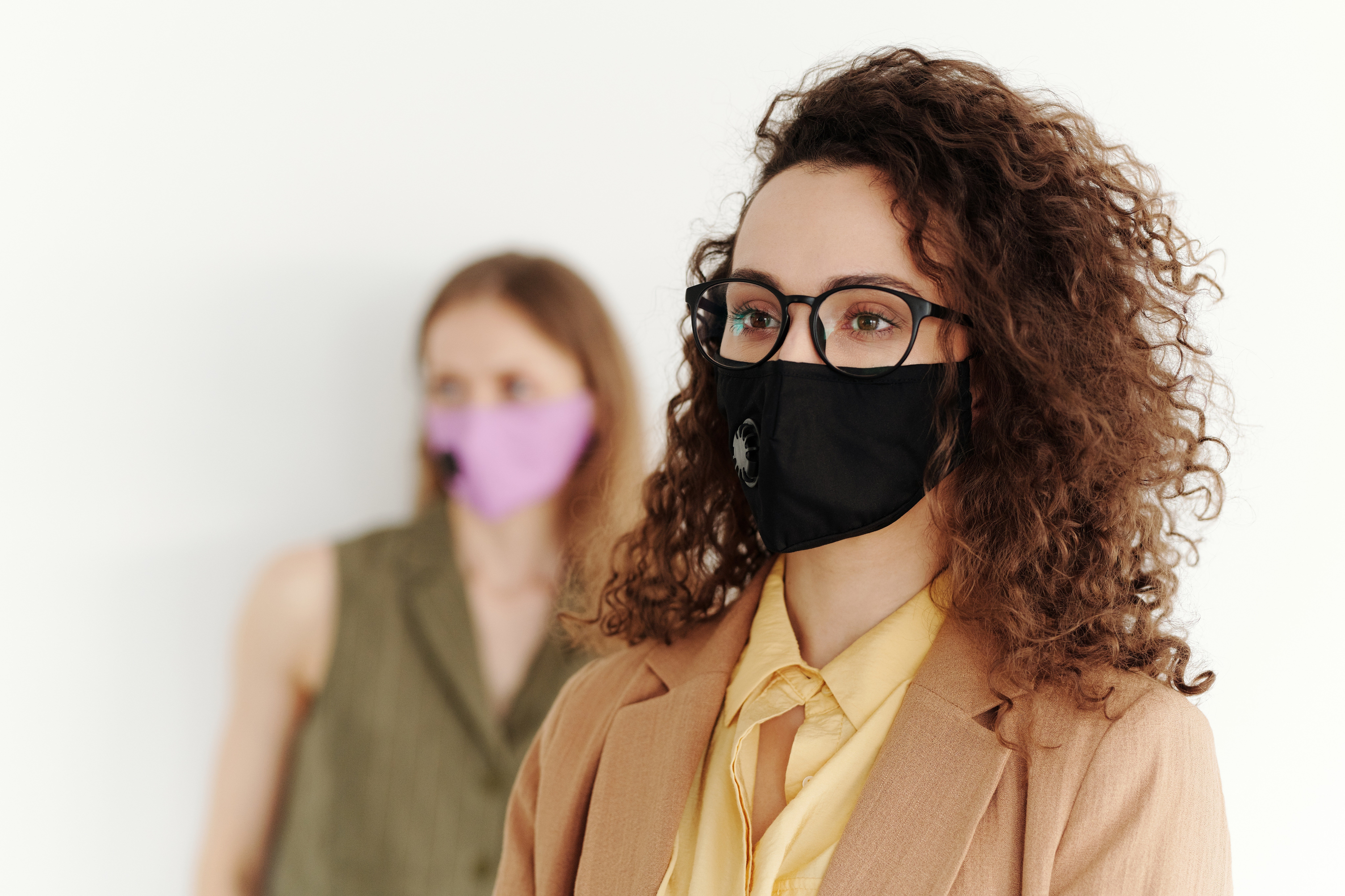 Two young businesswomen wearing cloth face masks look away from the camera.