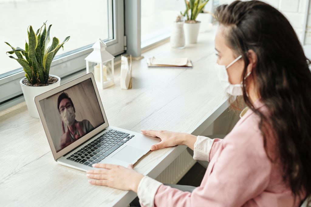 a young Caucasian woman in an office wearing a face mask looks into her laptop on a video conference with a young Asian coworker also wearing a mask.
