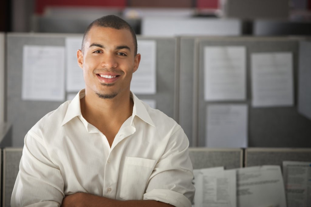 Young mixed-race business-casual man facing camera smiling with arms crossed with cubicles in background
