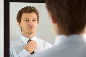 Photo of a younger white man looking determinedly into a mirror at himself and adjusting his tie.