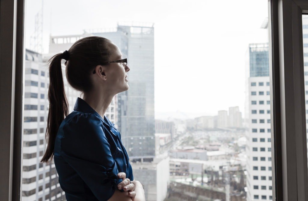 Young white woman in business casual staring pensively out the window of an office building