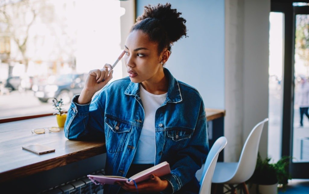 Thoughtful young black woman sitting in cafe with textbook in hand and thinking about plans for future